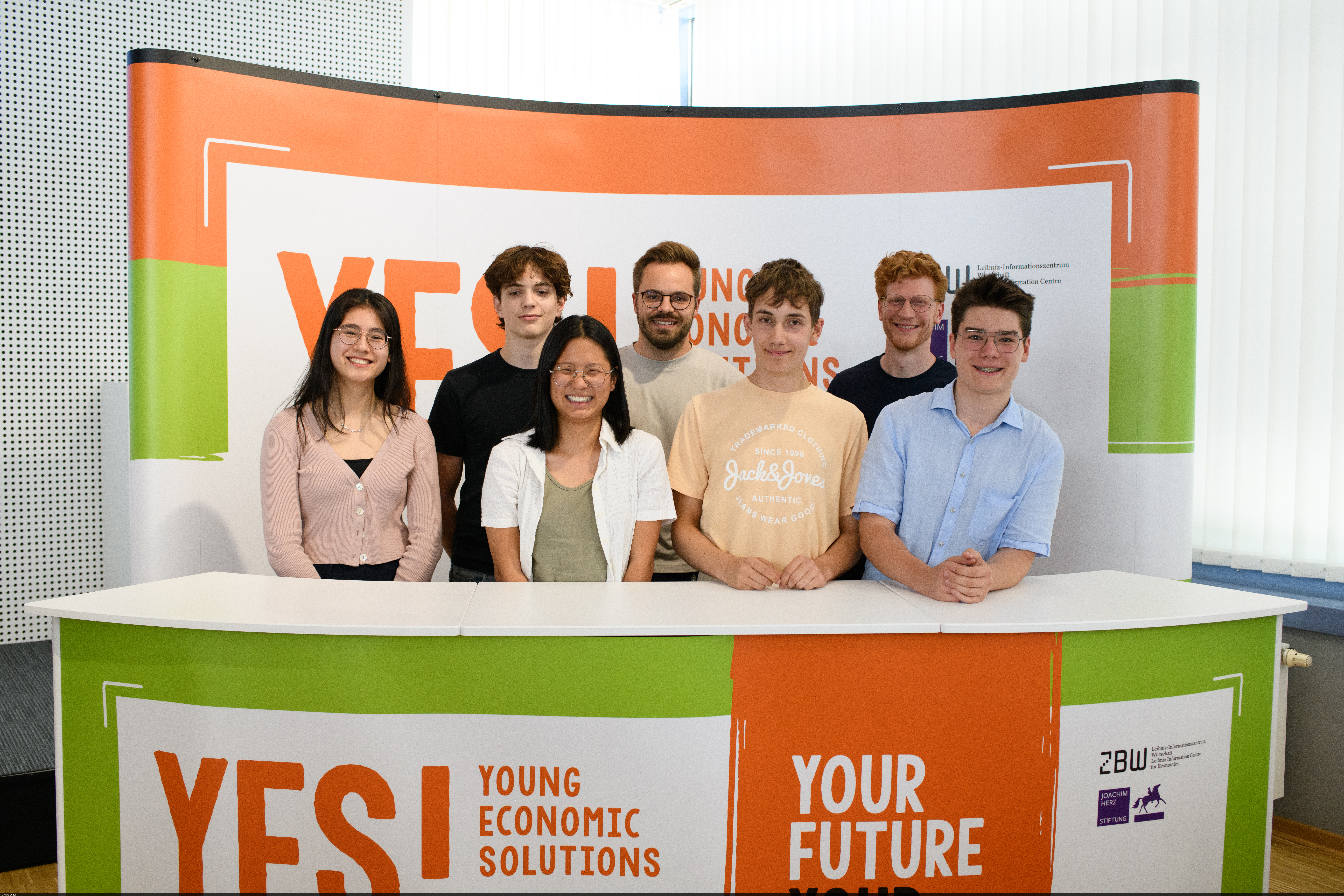 Schülerinnen und Schüler stehen hinter einem Pult mit einem „YES! Economic Solutions“-Banner.