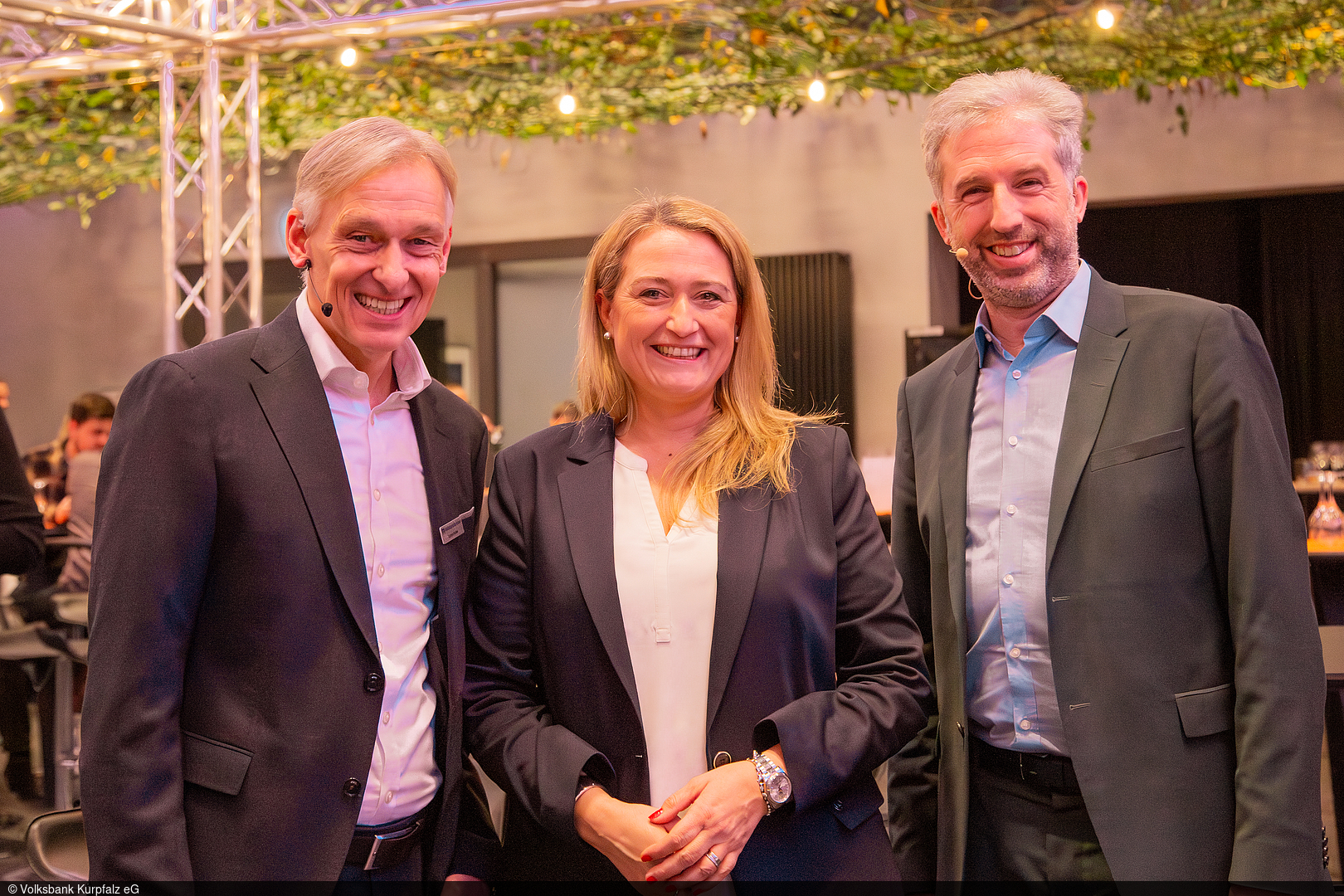 Drei lächelnde Erwachsene in Businesskleidung stehen nebeneinander bei einer Veranstaltung. Alle tragen Ansteckmikrofone. Im Hintergrund sind Sitzplätze, eine Bühne mit Metallträgern sowie dekorative Lichter und Pflanzen zu sehen.