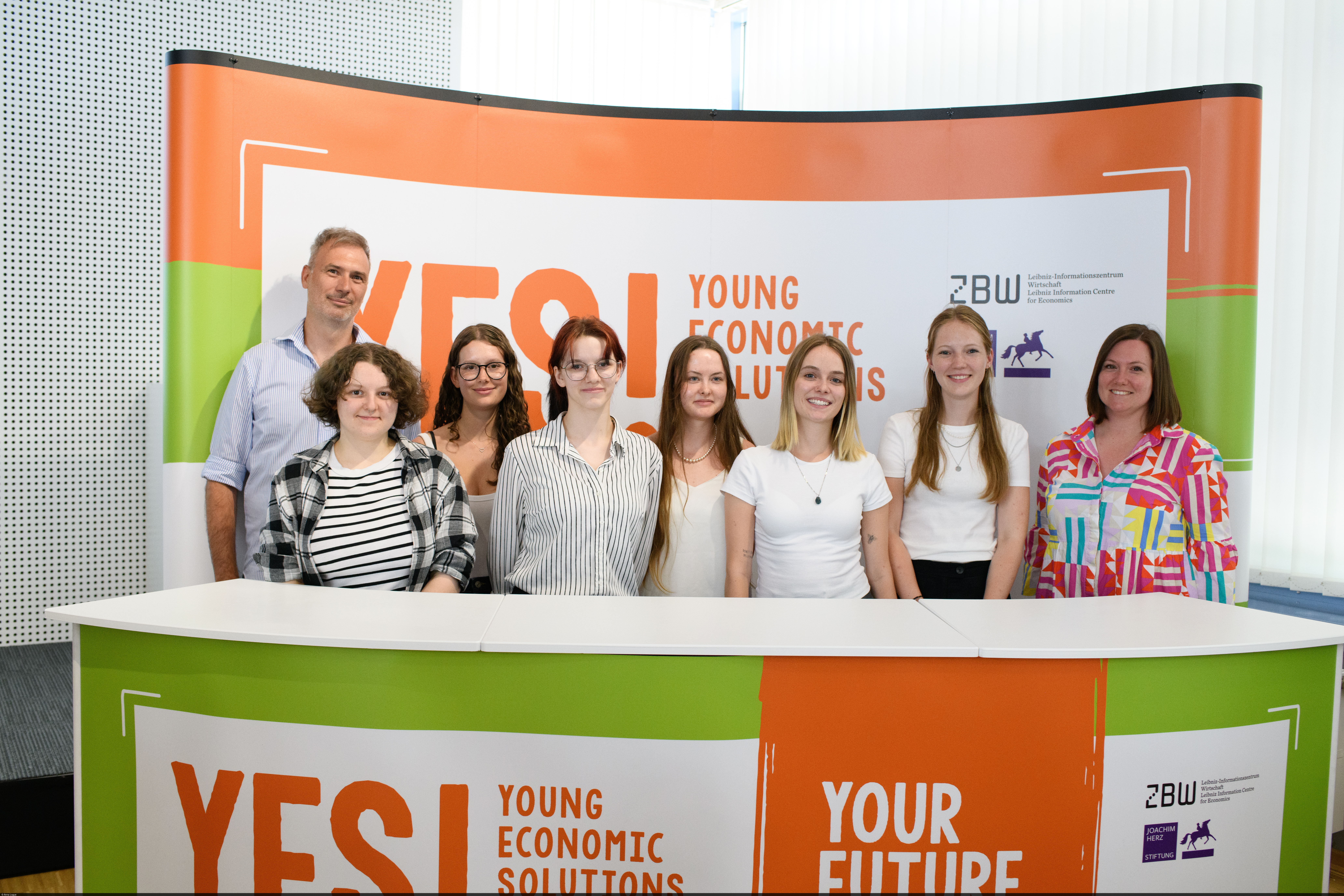 Schülerinnen und Schüler stehen hinter einem Pult mit einem „YES! Economic Solutions“-Banner.