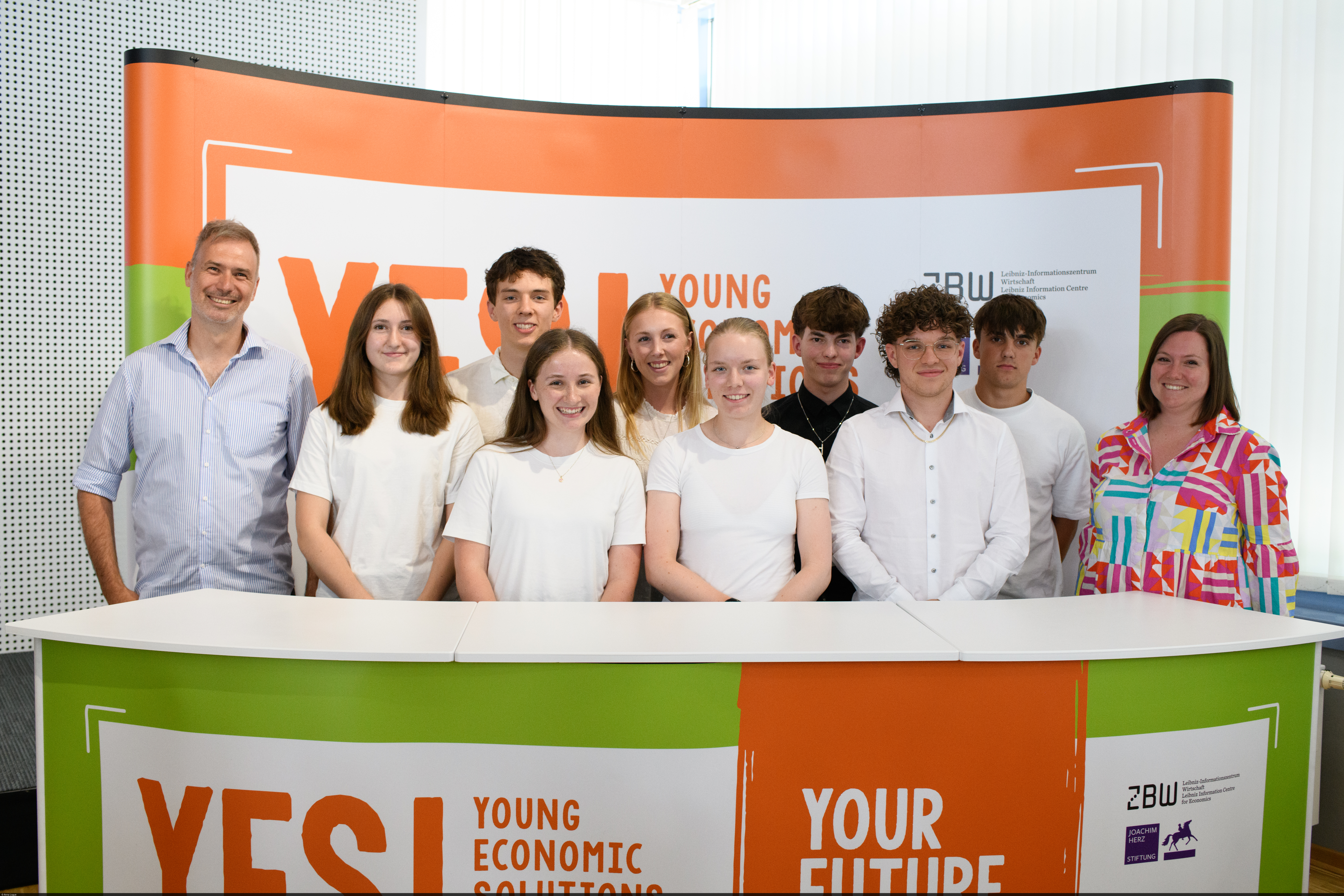 Schülerinnen und Schüler stehen hinter einem Pult mit einem „YES! Economic Solutions“-Banner.