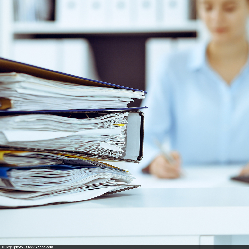 Image of a woman at a table processing documents. Three full file folders lie to her left in the foreground of the image.