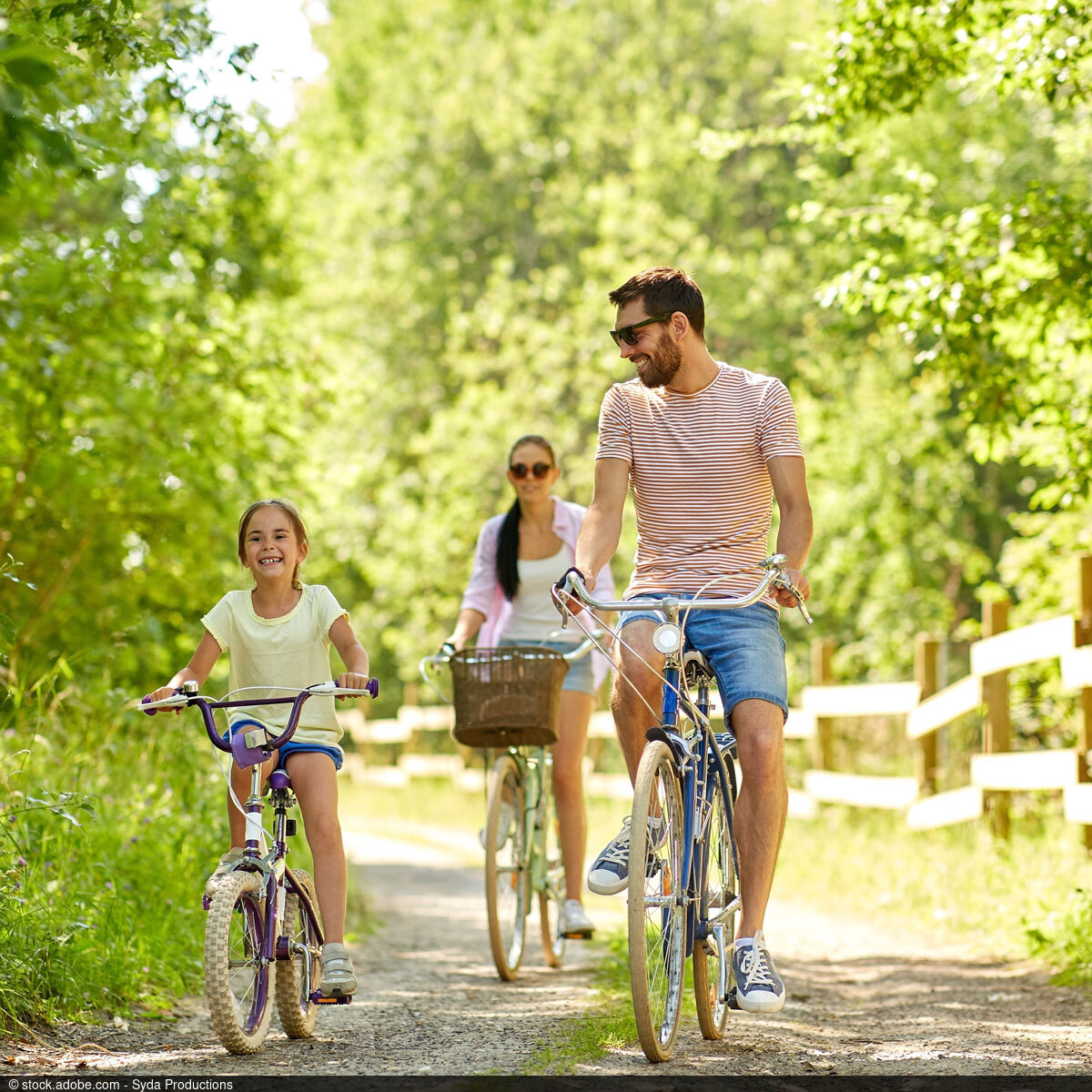 Lächelndes Mädchen fährt auf einem Waldweg Fahrrad, neben ihr ein Mann; im Hintergrund fährt eine Frau an einem sonnigen Tag.
