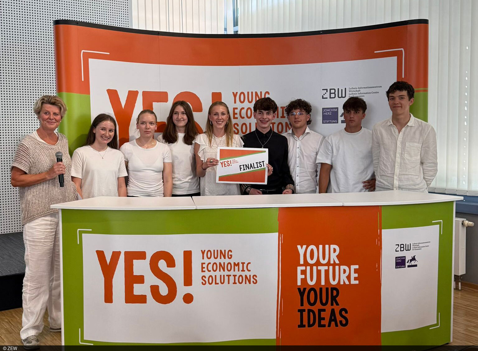 Schülerinnen und Schüler posieren hinter einem Pult mit einem „YES! Economic Solutions“-Banner.