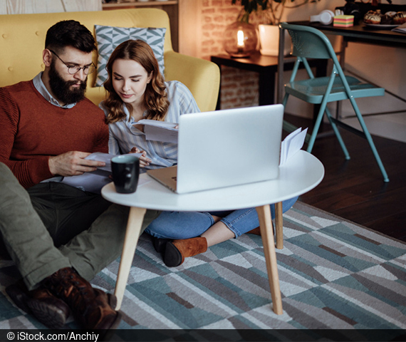 Ein Mann und eine Frau sitzen vor einem Laptop und vergleichen Papiere, die sie in den Händen halten.