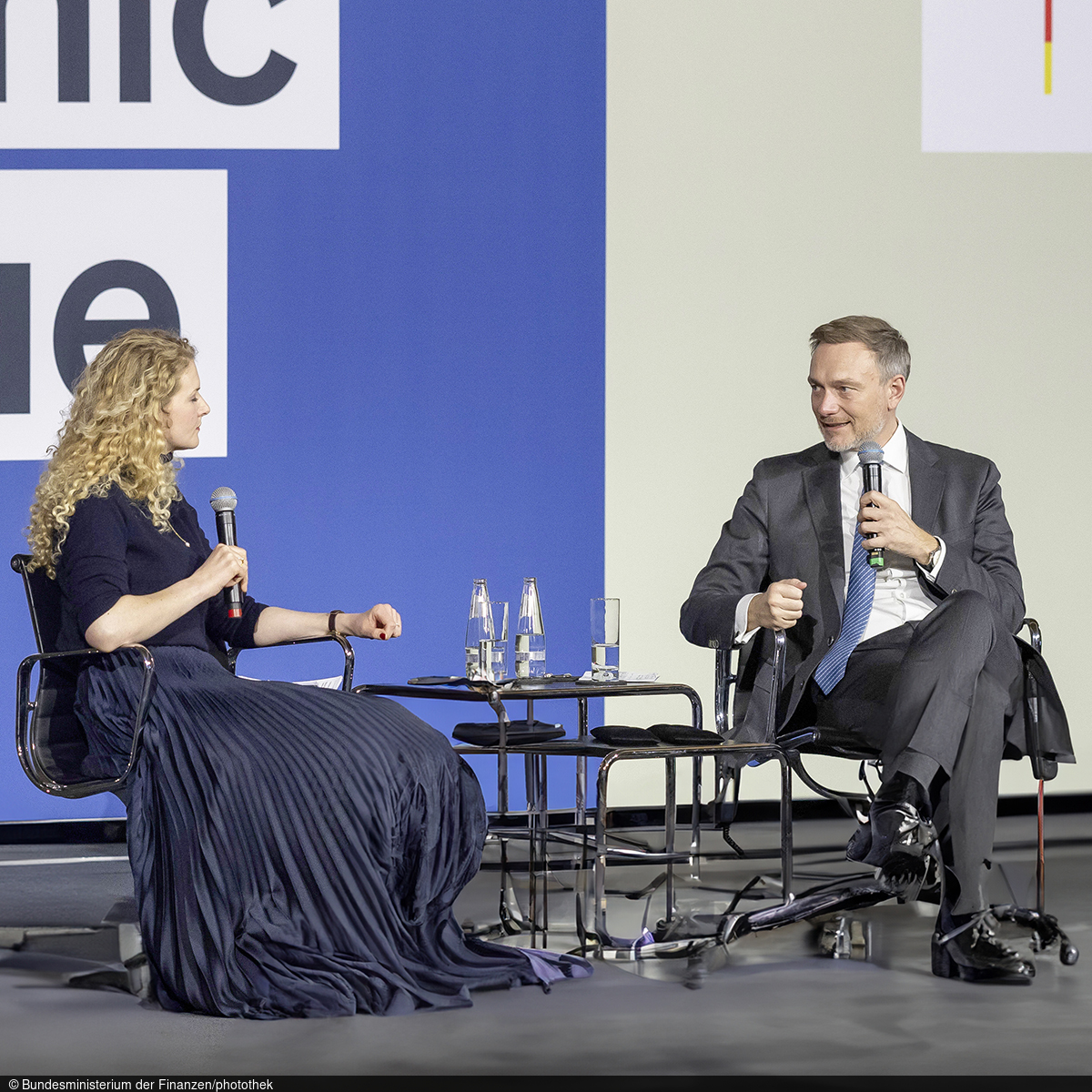 The presenter sits on a stage next to Federal Minister Christian Lindner at the Economic Dialogue