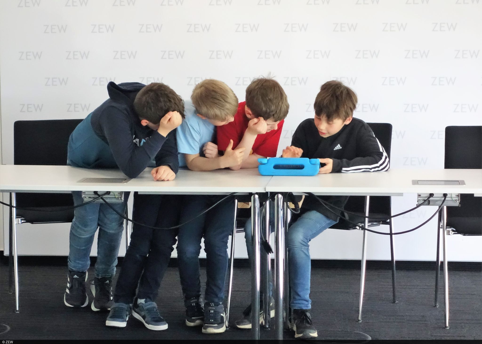 Kinder, die gemeinsam am Tisch sitzen/stehen und sich mit einem Tablet beschäftigen