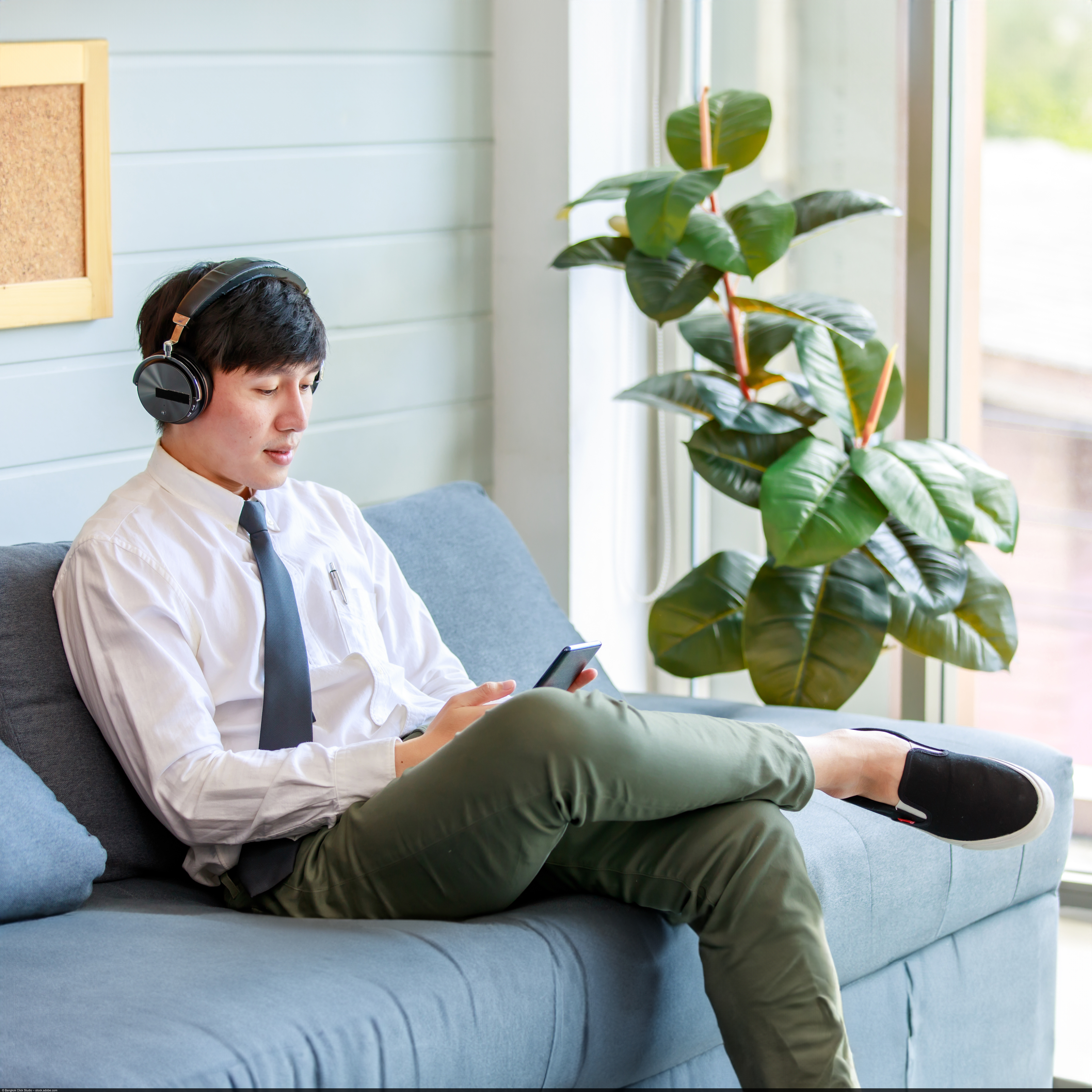 A young man sits on a couch and listens to music via smartphone and headphones. In the background is a green houseplant.