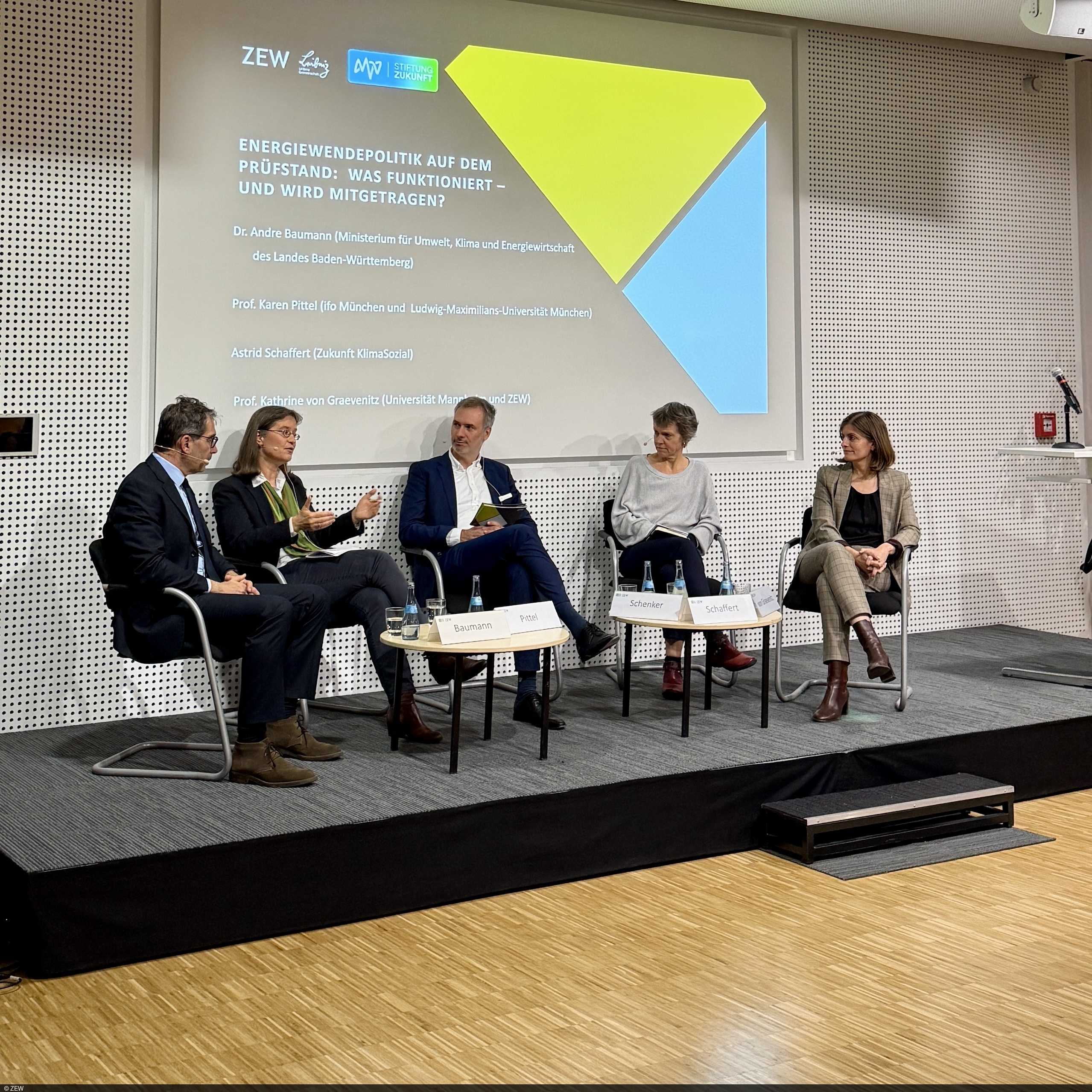 Eine Podiumsdiskussion mit fünf Personen auf einer Bühne. Vier Frauen und ein Mann sitzen in einer Reihe auf modernen Stühlen. Hinter ihnen befindet sich eine große Projektionsfläche mit einer farbigen Grafik und Text zur Energiewendepolitik. Die Teilnehmenden sprechen miteinander, einige halten Notizen in der Hand. Vor ihnen stehen kleine Tische mit Wasserflaschen und Namensschildern. Der Raum wirkt professionell ausgestattet, mit hellen Wänden und einem Holzboden.