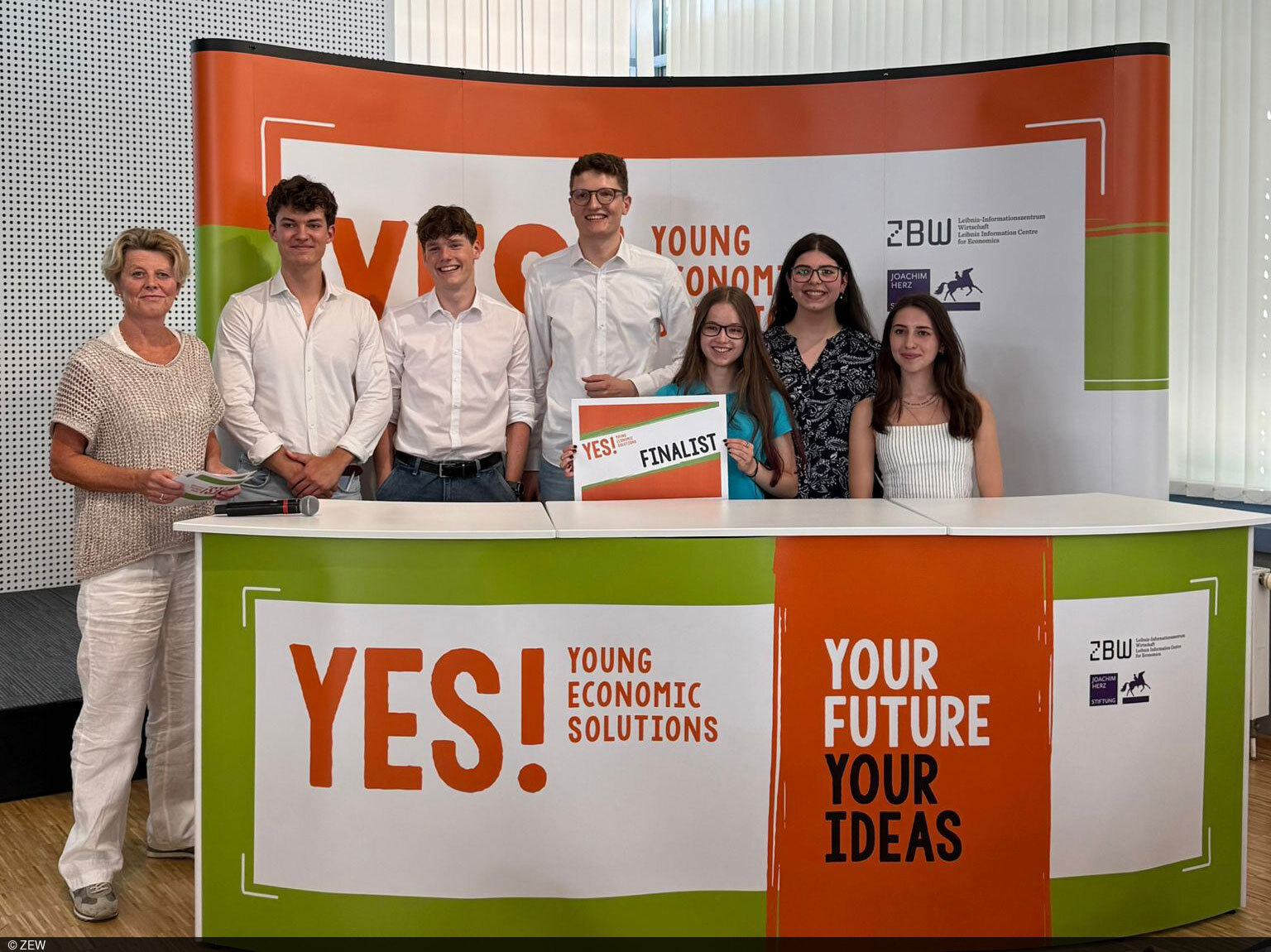 Schülerinnen und Schüler stehen hinter einem Pult mit einem „YES! Economic Solutions“-Banner.
