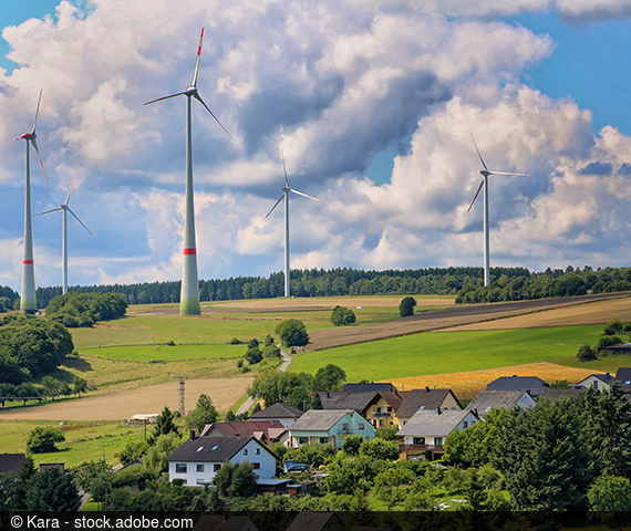 Photo of a village in the green with wind turbines in the background.