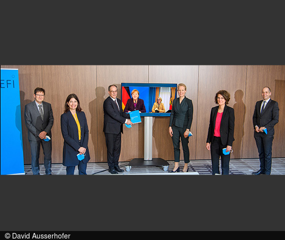 Die EFI-Mitglieder stehen um einen Bildschirm, auf dem Bundeskanzlerin Merkel und Bundesforschungsministerin Karliczek zu sehen sind.