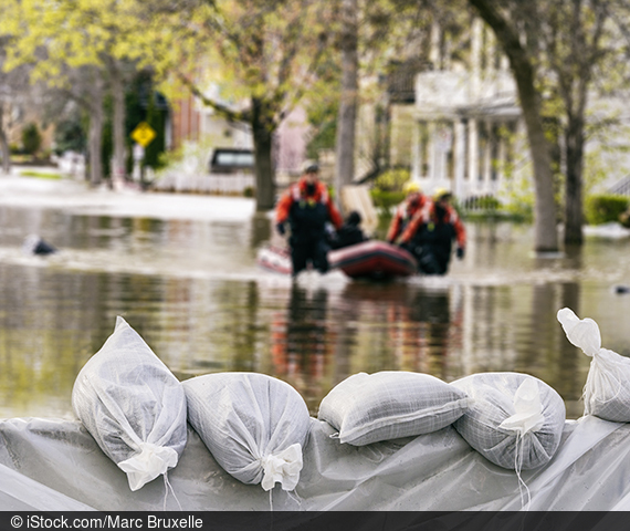 Sandbags hold back flood waters. Behind them, helpers wade through floodwaters in a city and push a blue boat