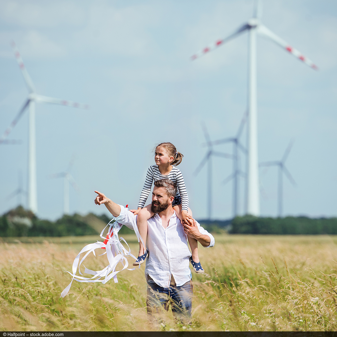 Vater läuft mit Tochter über ein Feld. Im Hintergrund befinden sich Windräder.