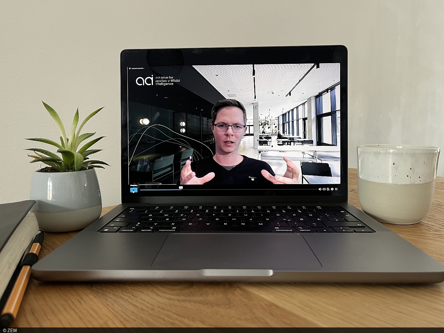 There is a laptop on a wooden table, on whose screen Andreas Liebl can be seen talking.