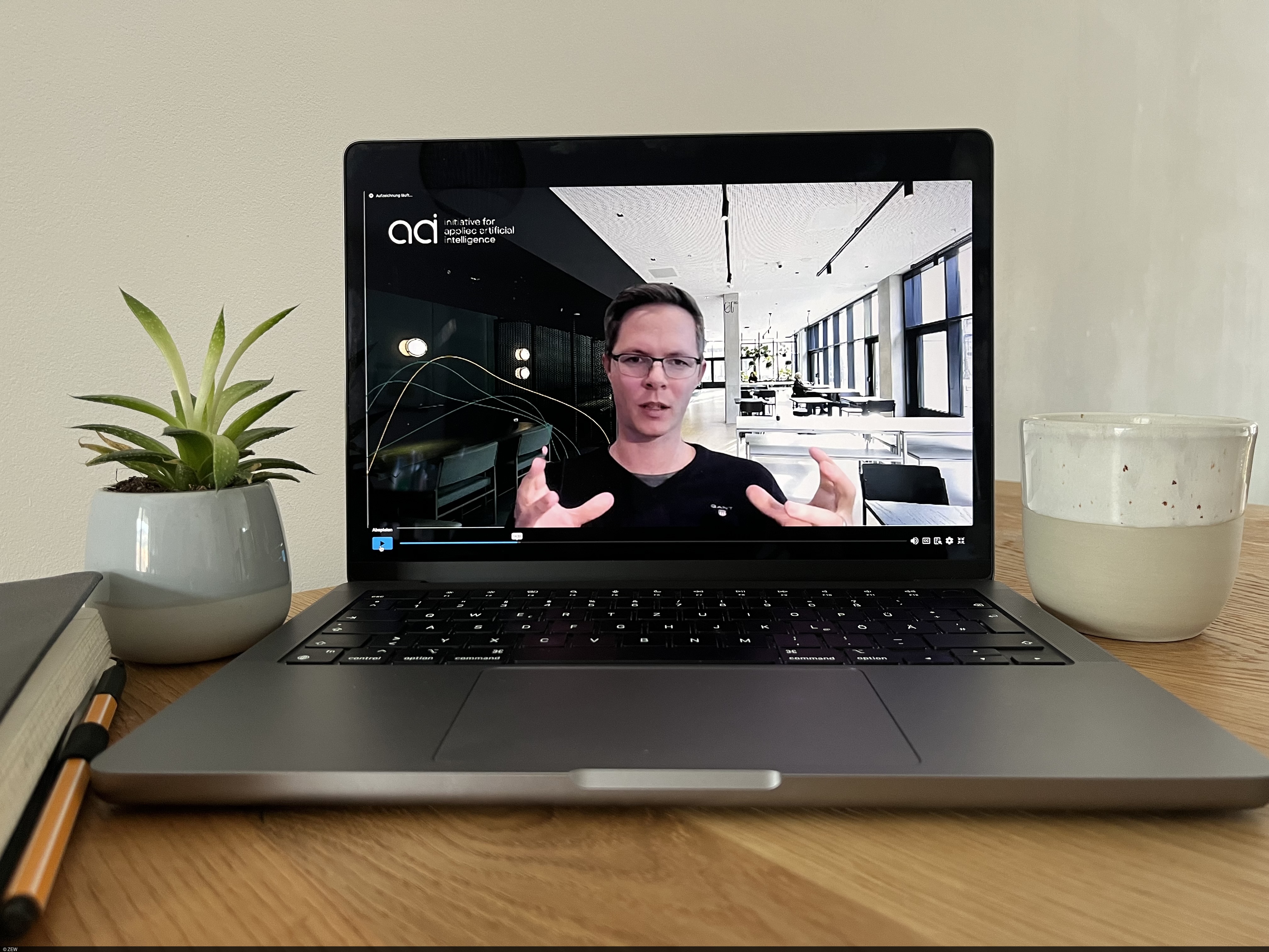 Auf einem Holztisch steht ein Laptop, auf dessen Bildschirm ist Andreas Liebl im Vortrag zu sehen.