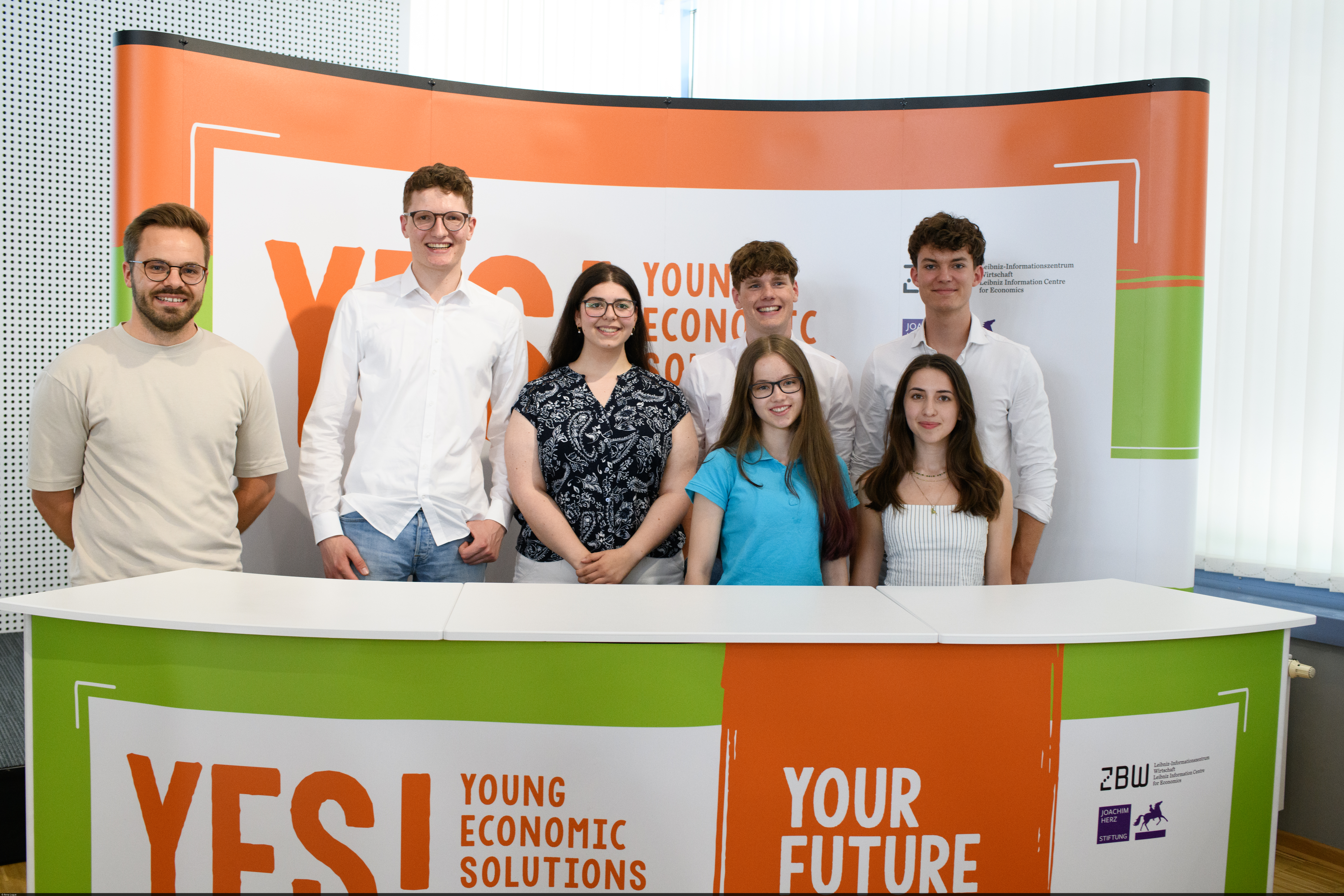 Schülerinnen und Schüler stehen hinter einem Pult mit einem „YES! Economic Solutions“-Banner.